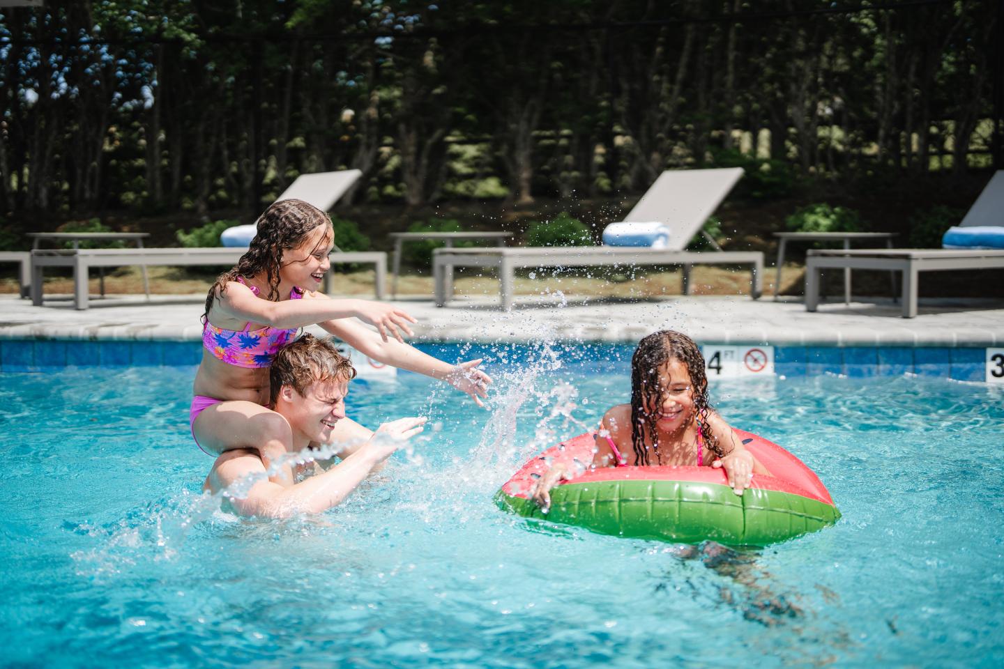 Kids playing in a pool, one on a float, others splashing water.