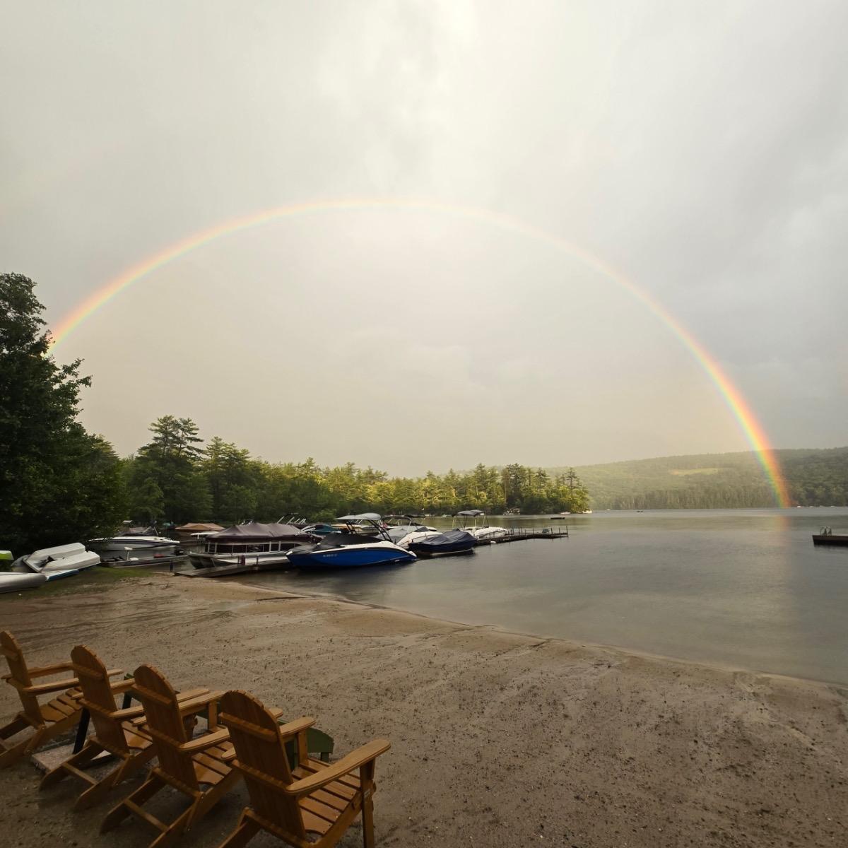 Sandy lakeside with chairs, boats docked, and a rainbow in the cloudy sky.