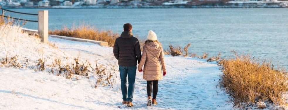 Couple walking on snowy path by a lake, dressed in winter clothes.
