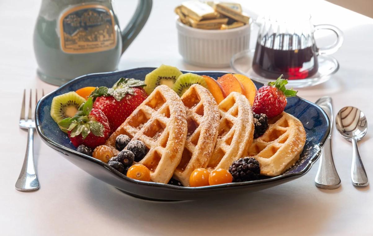 Waffles with fruit on a blue plate, with syrup and a green jug nearby.