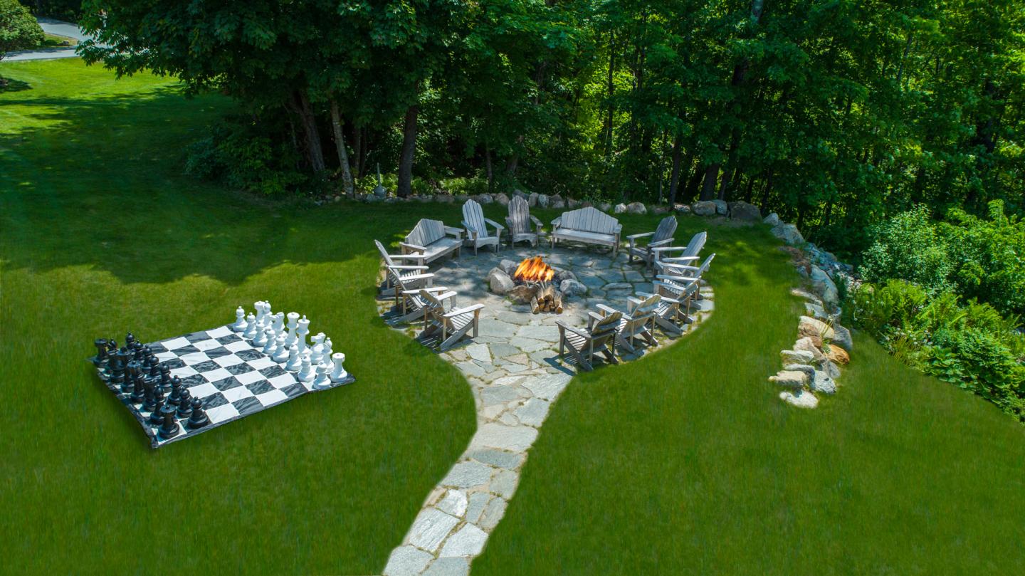 Stone path leads to a fire pit with chairs, surrounded by trees; large chessboard nearby.