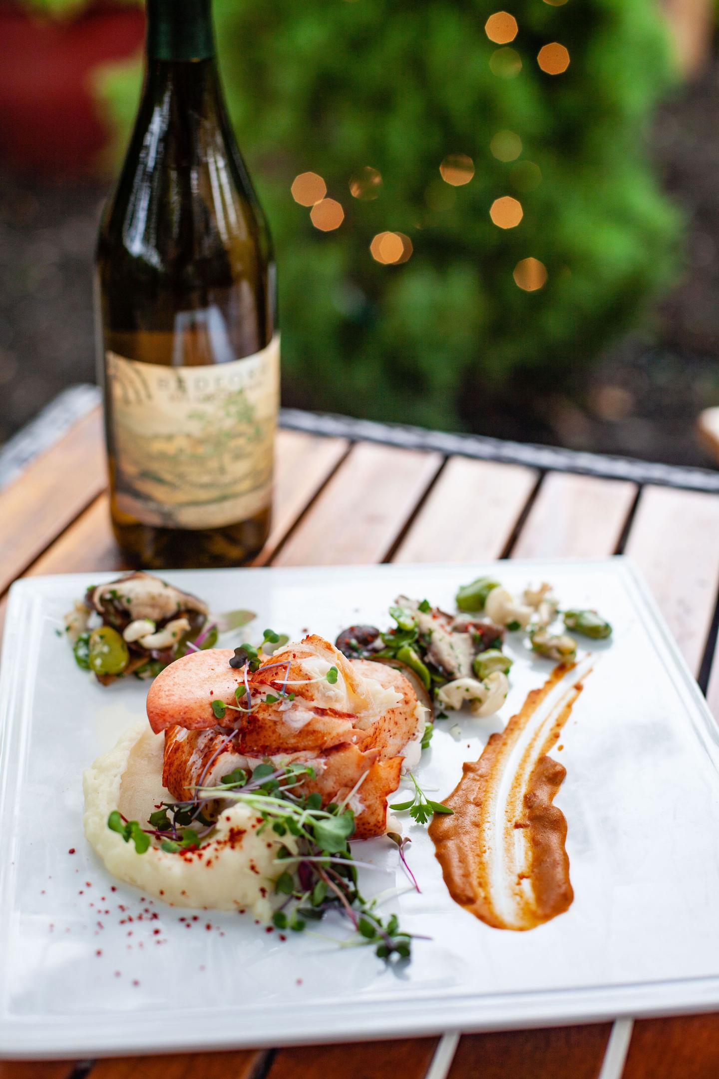 Gourmet dish with mashed potatoes, greens, and salmon on a square plate, wine bottle nearby.