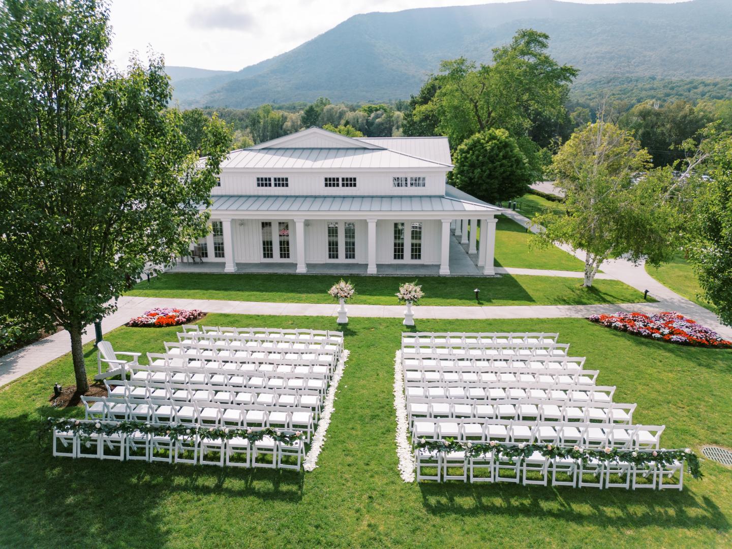 White chairs arranged for an outdoor event facing a large white house with mountains in the background.