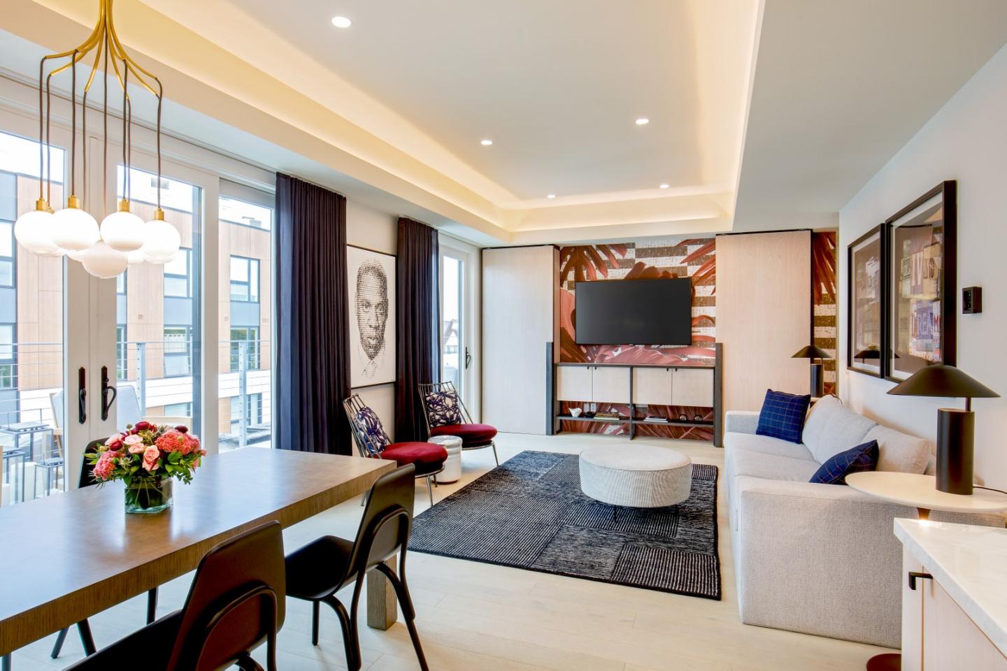 Modern living room with large windows, a dining table, and a wall-mounted TV.