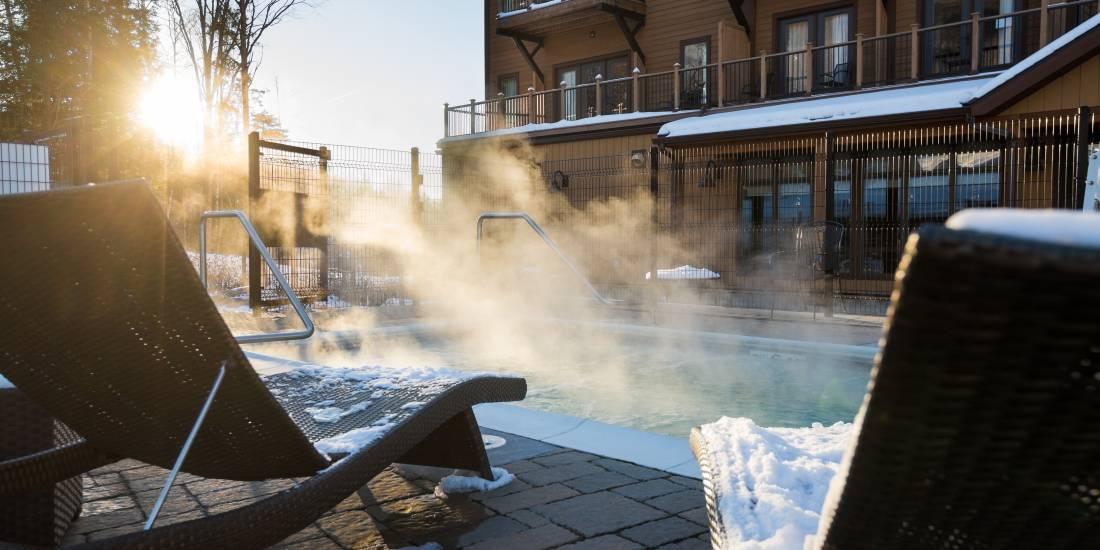 Sunlit outdoor pool with steam rising, snowy surroundings, and lounge chairs.