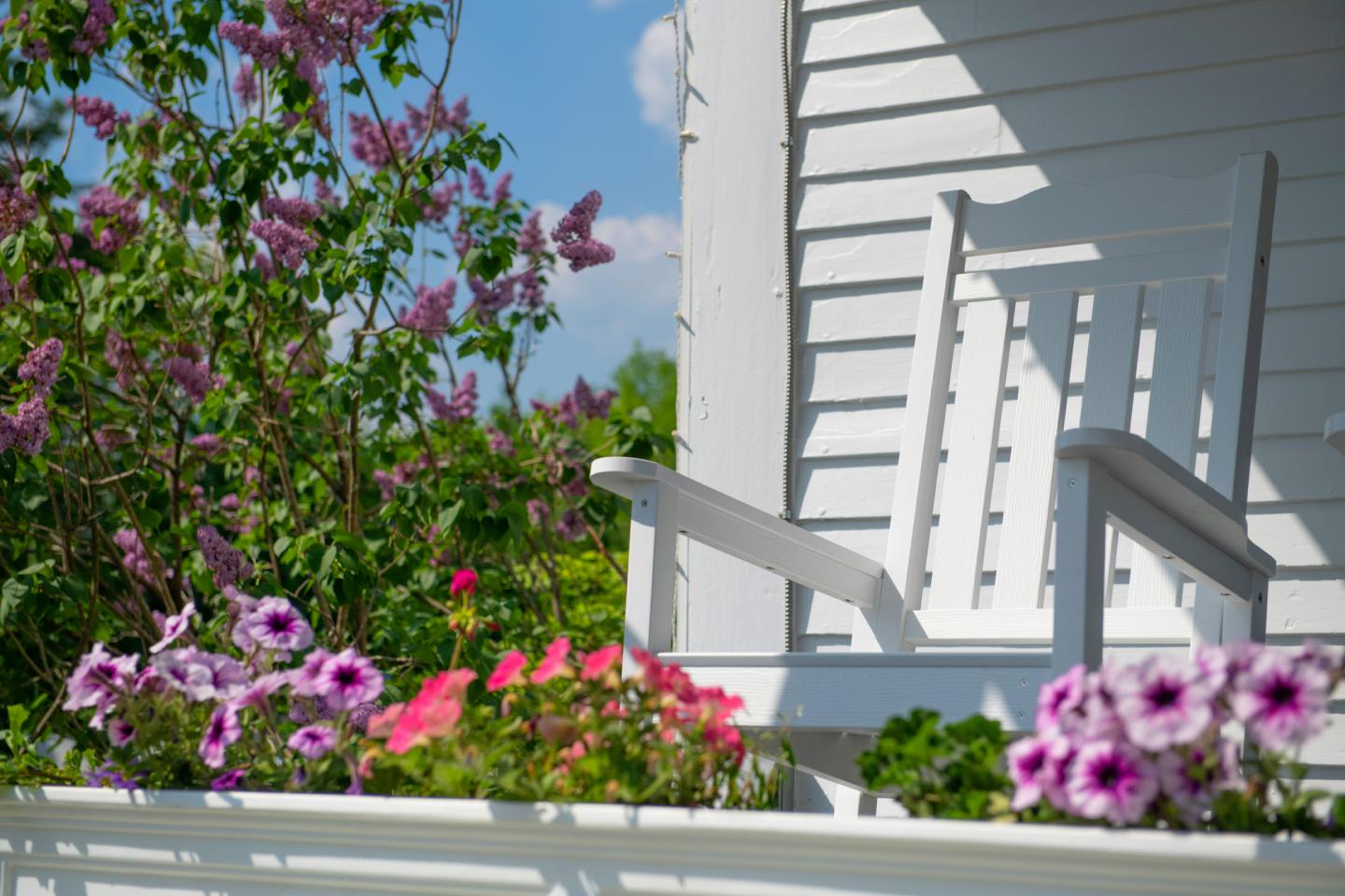 White porch with rocking chair, flowers, and purple bushes under a blue sky.