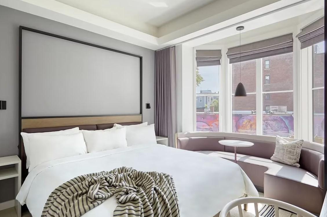 Modern hotel room with a large bed, corner seating, and large windows.