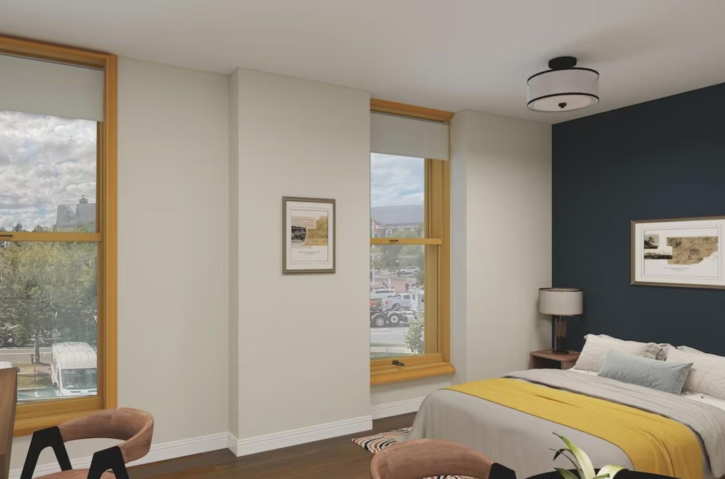 Bedroom with large windows, a bed with yellow blanket, and framed artwork on walls.