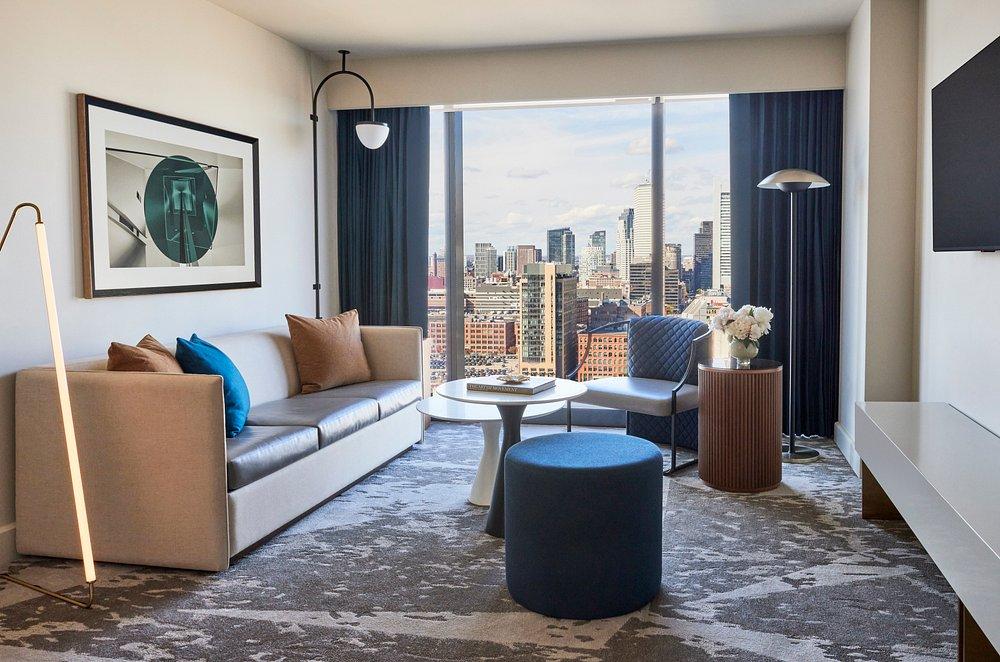 Modern hotel room with city view, sofa, blue ottoman, and chairs.