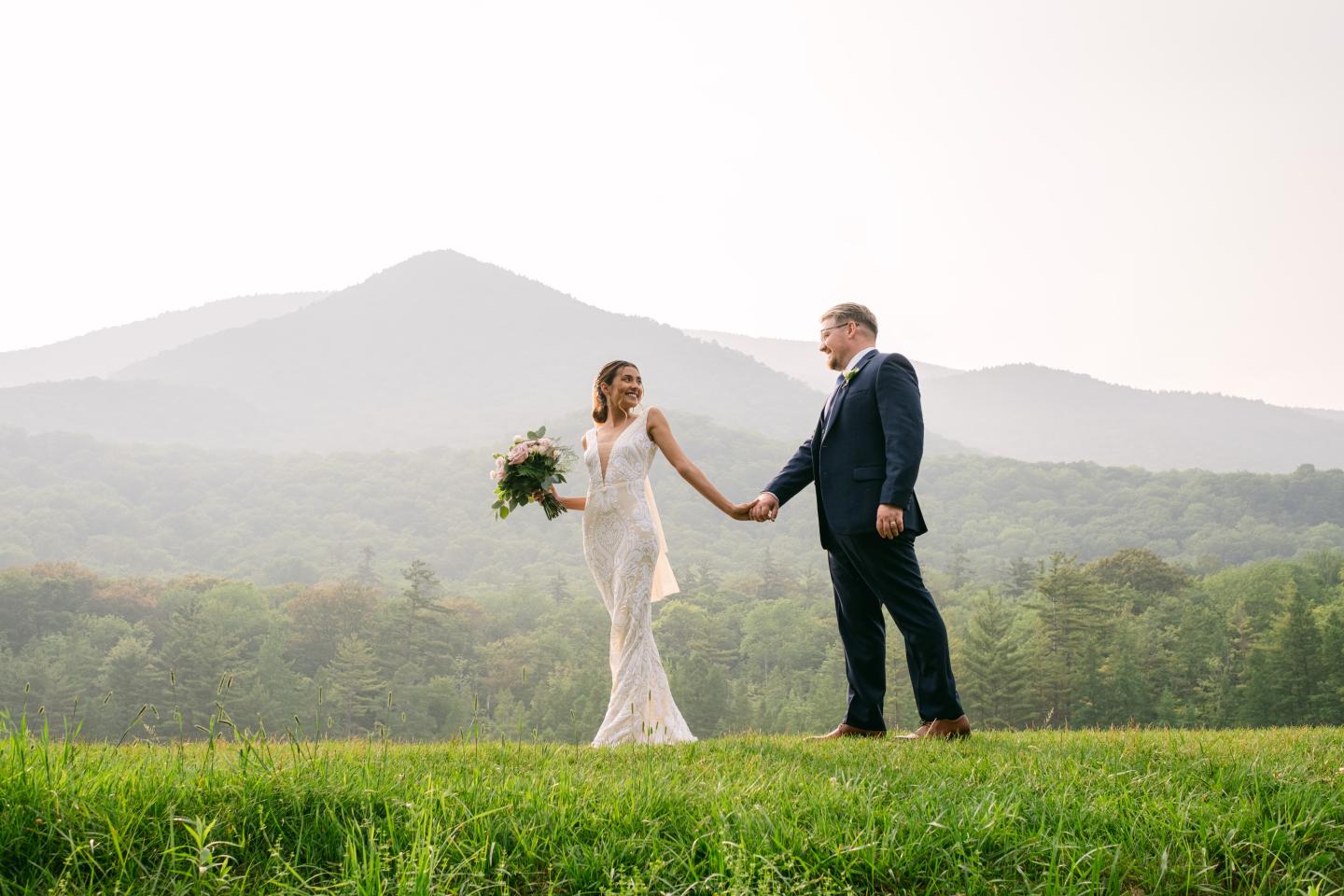 Bride and groom holding hands on a grassy hill, mountains in the background.