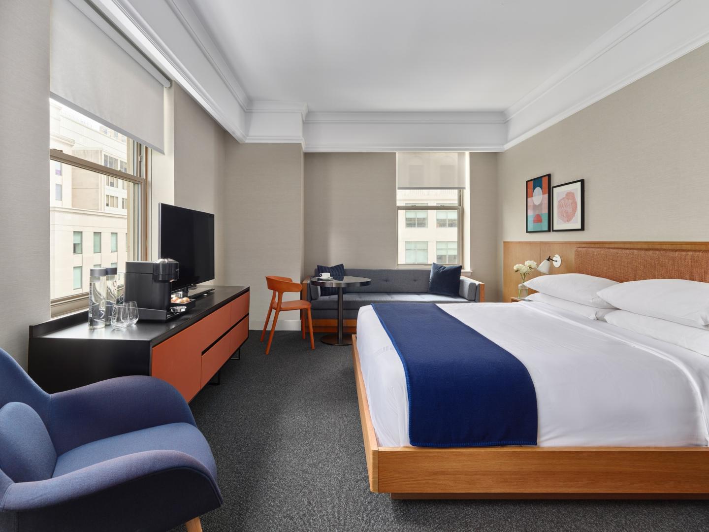 Modern hotel room with a king bed, TV, armchair, and large windows.
