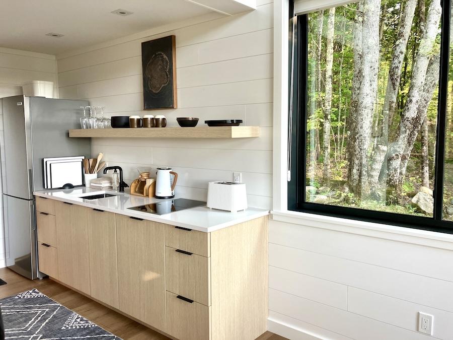 Modern kitchen with light wood cabinets, shelf, and large window facing trees.