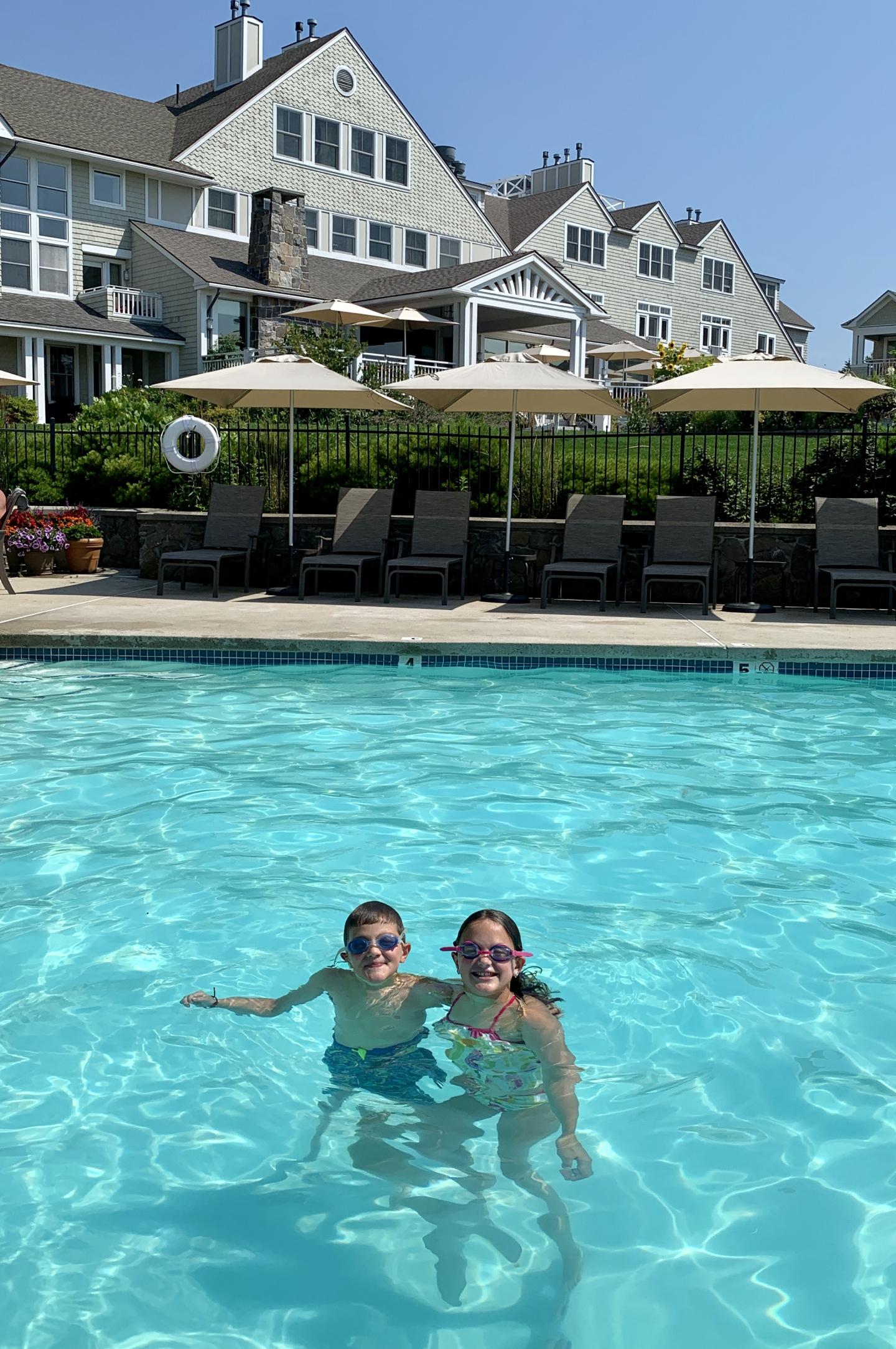 Two people smiling in a pool, with houses and umbrellas in the background on a sunny day.