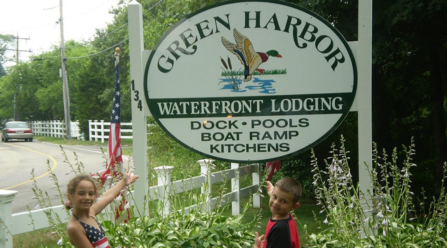 Kids smiling and pointing at Green Harbor Lodging sign by the roadside.