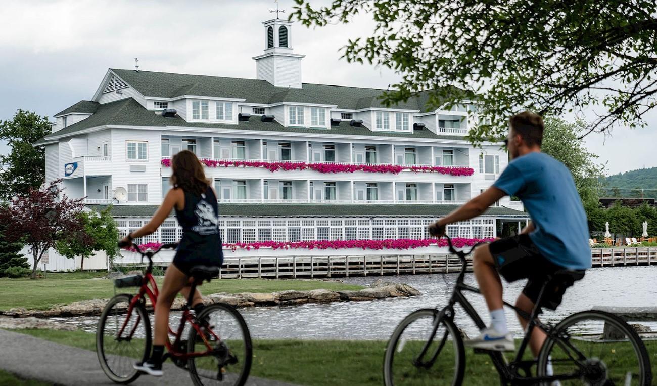 Cyclists ride past a large white lakeside building.