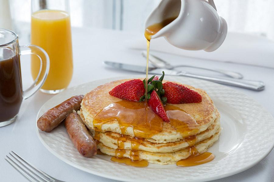 Pancakes with syrup, strawberries, and sausages, next to coffee and orange juice.