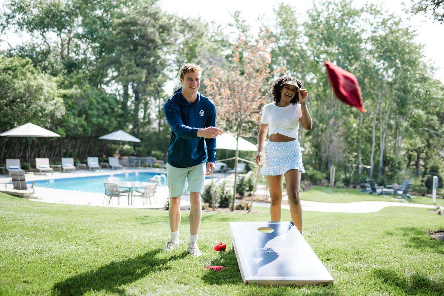 Two people playing cornhole near a pool on a sunny day.