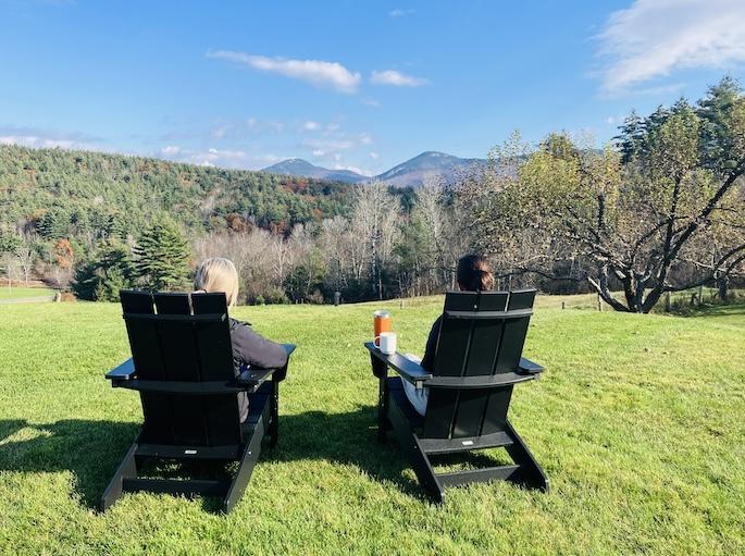 Two people sit in chairs on a grassy hill, overlooking mountains and trees.