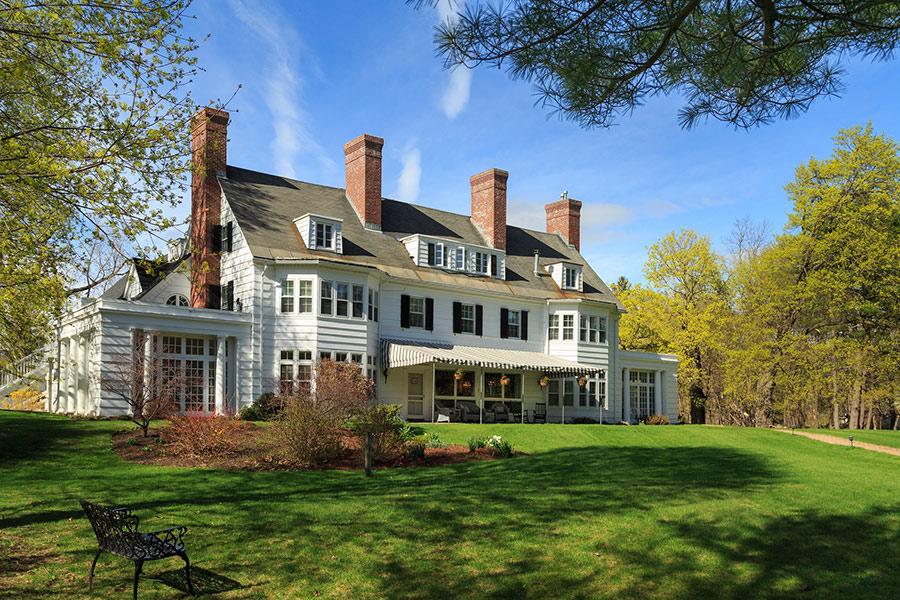 Large white house with four chimneys, set in a green lawn under a blue sky.