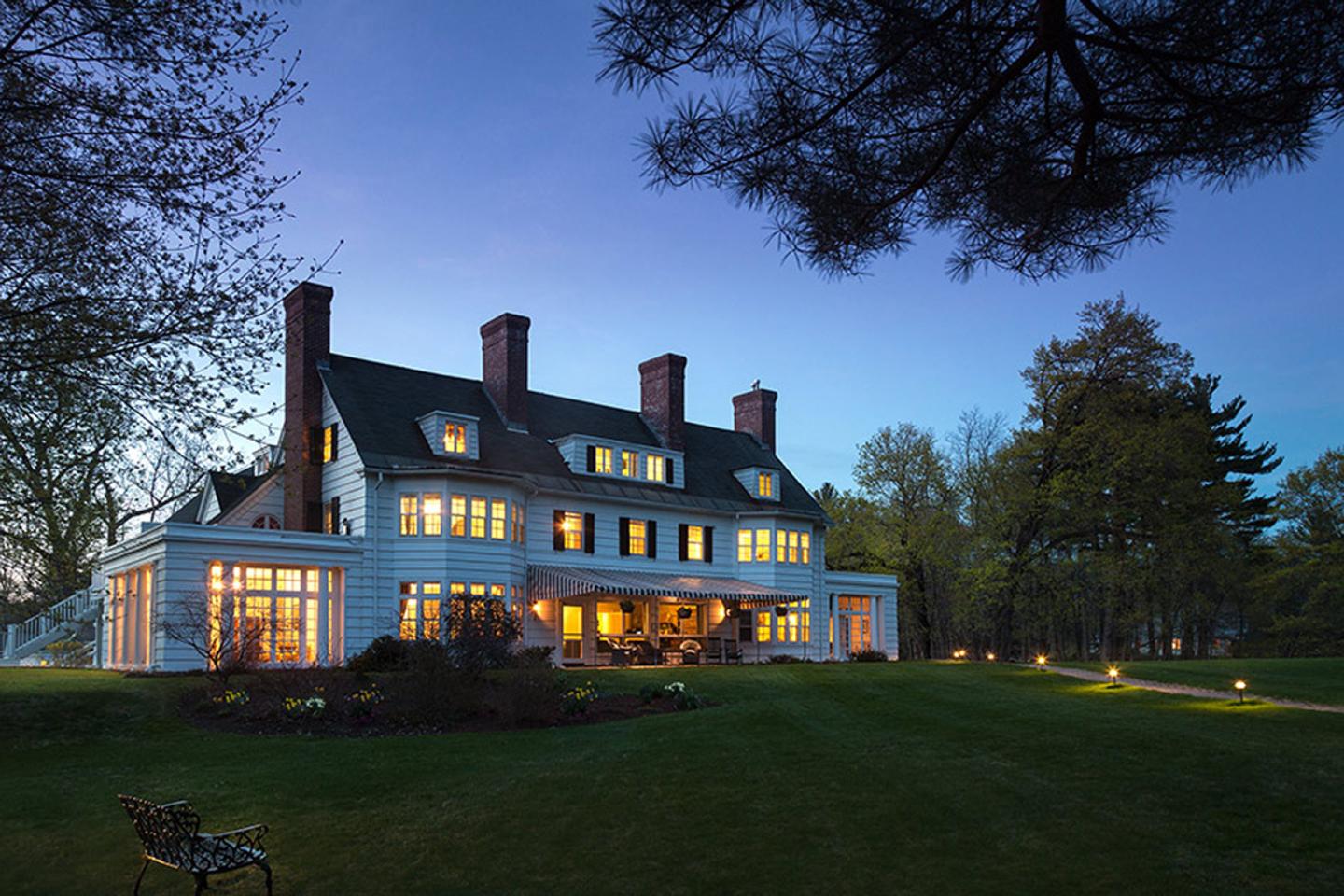 Large house with lit windows at dusk, surrounded by trees and a spacious lawn.