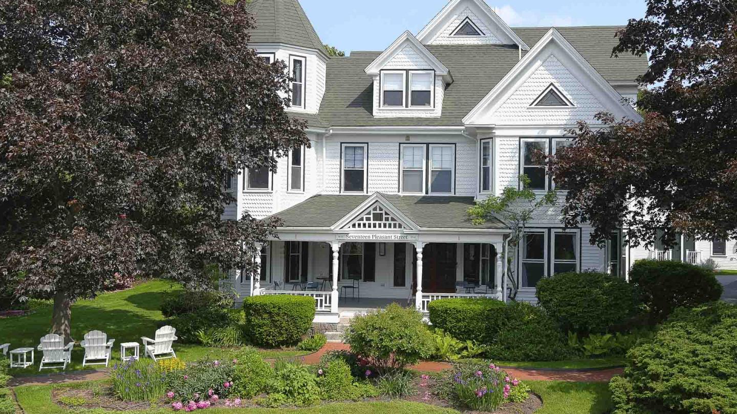 Large white Victorian house with a porch, surrounded by trees and a garden.