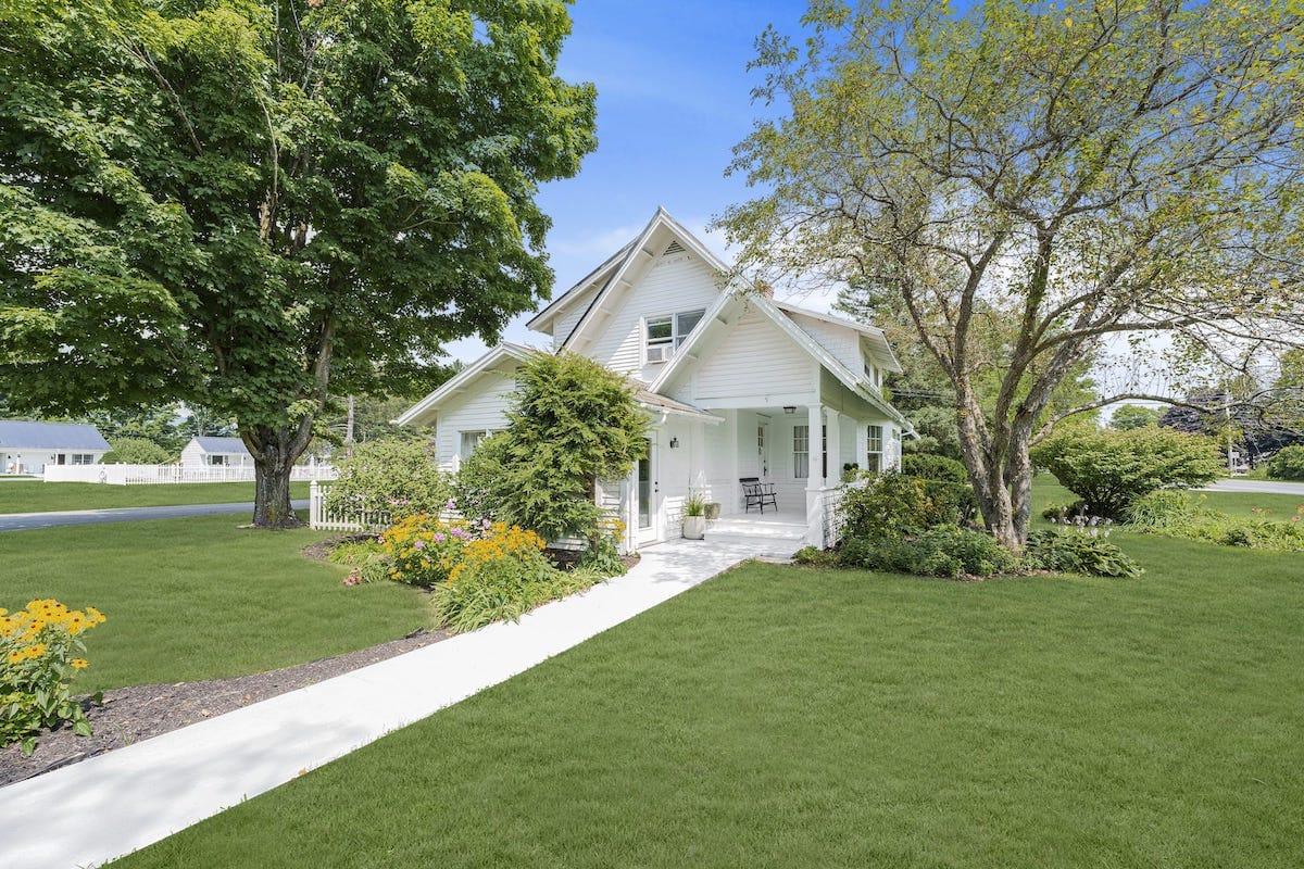 Charming white house with porch, surrounded by lush garden and trees.