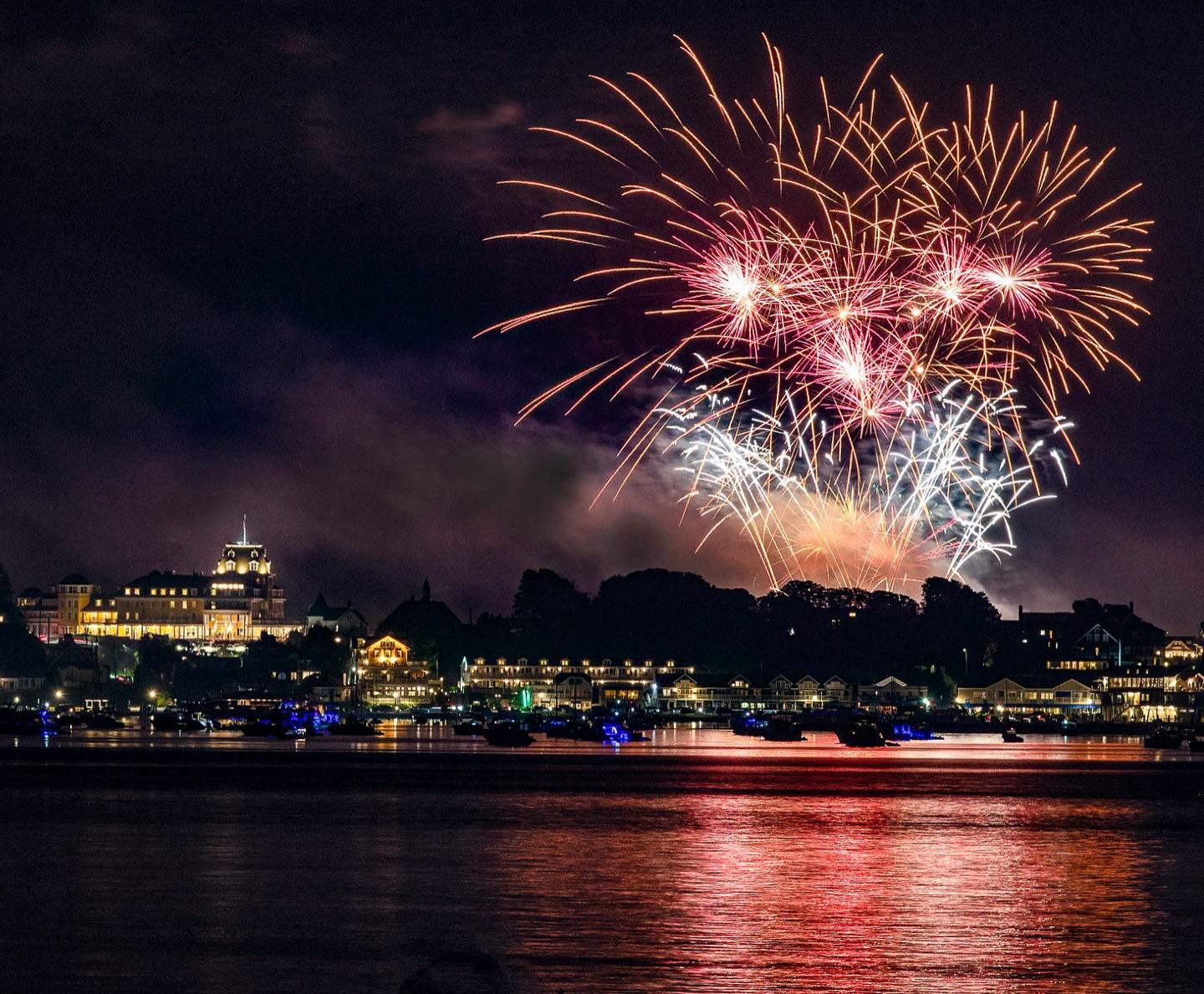 Fireworks burst over a city skyline at night, reflecting on the water.