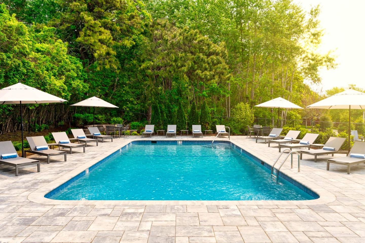 Outdoor pool with lounge chairs and umbrellas, surrounded by lush green trees.