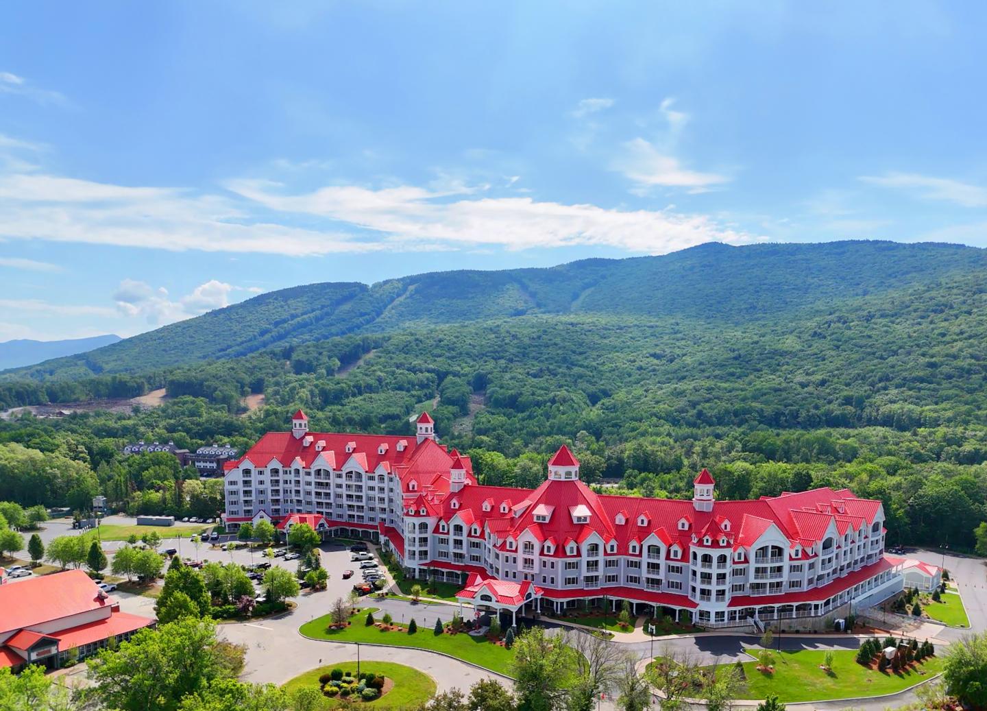 Large hotel with red roofs, surrounded by green mountains under a blue sky.
