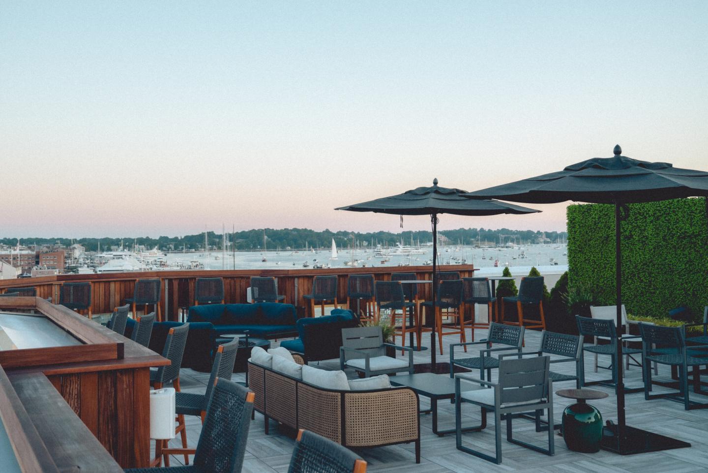 Rooftop patio with tables, chairs, and umbrellas overlooking a marina at sunset.