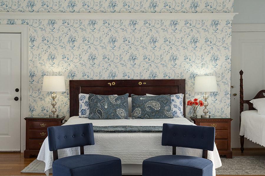 Cozy bedroom with floral wallpaper, blue chairs, dark wood bed, and matching nightstands.
