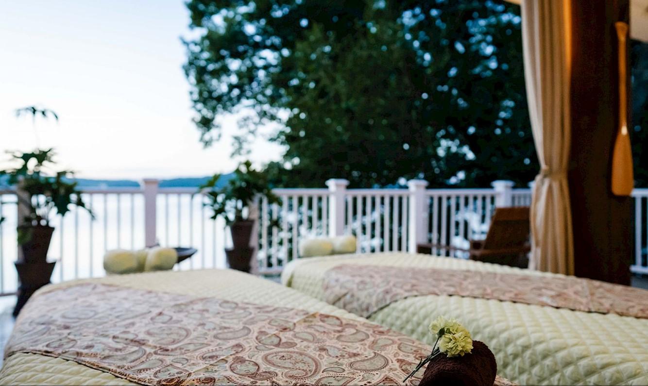 Two massage tables on a serene outdoor balcony with a lake view.