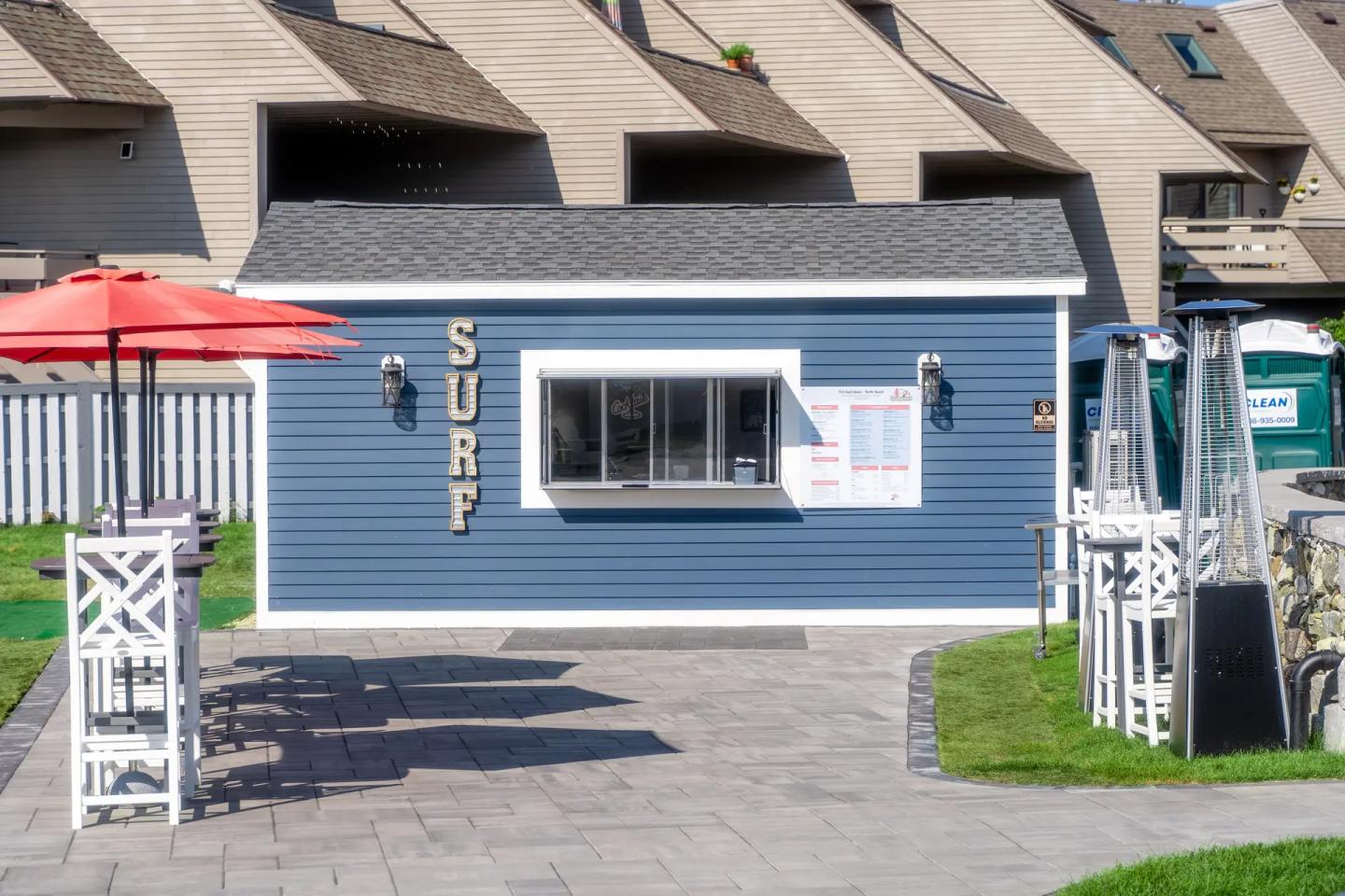 Blue surf shop with large window, red umbrellas, and modern wicker furniture.