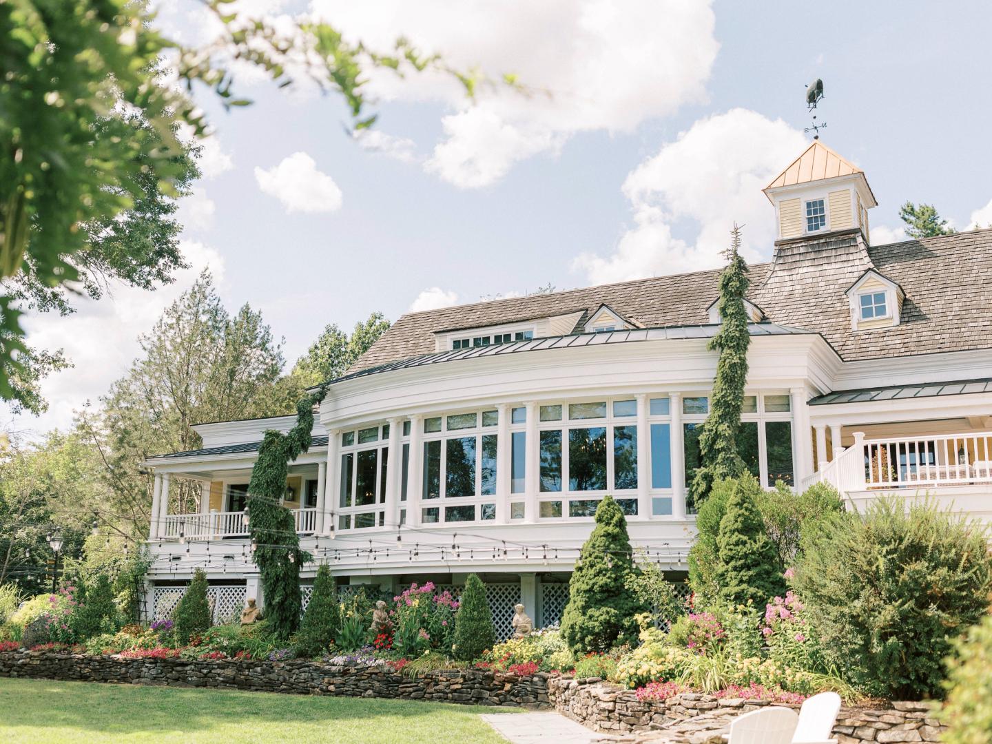Elegant white house with wraparound porch, surrounded by lush gardens under a blue sky.