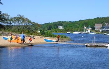 People kayaking near a sandy riverbank with trees and houses in the background.