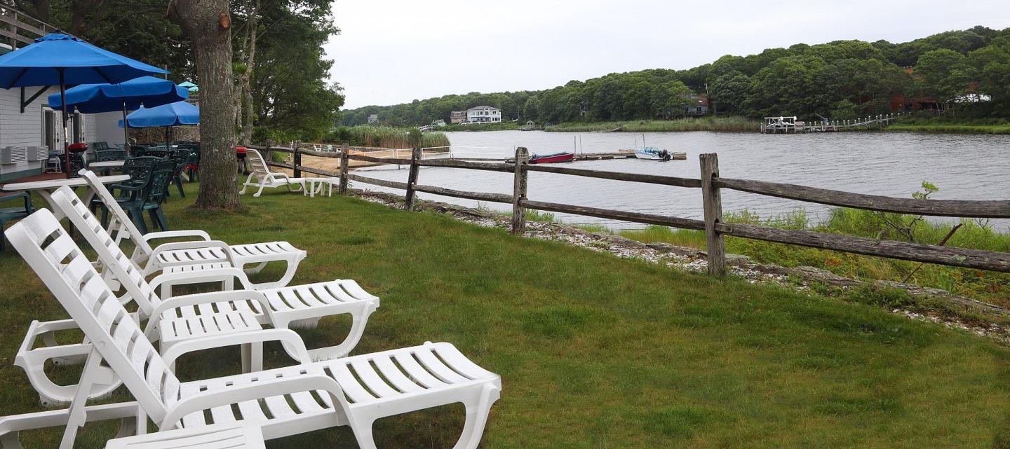 Row of white lounge chairs by a grassy riverbank with trees and overcast sky.