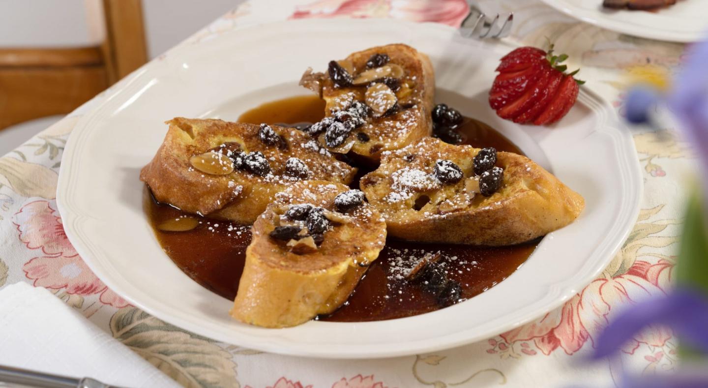 French toast with raisins, drizzled syrup, and a strawberry garnish on a floral tablecloth.