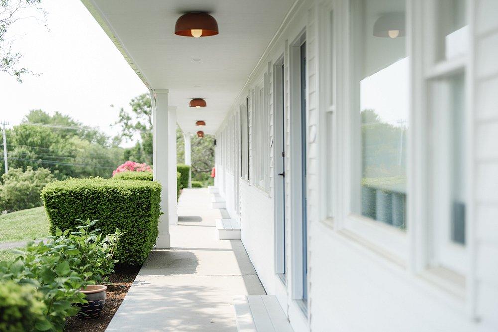 Motel corridor with white walls, red lights, and green bushes.