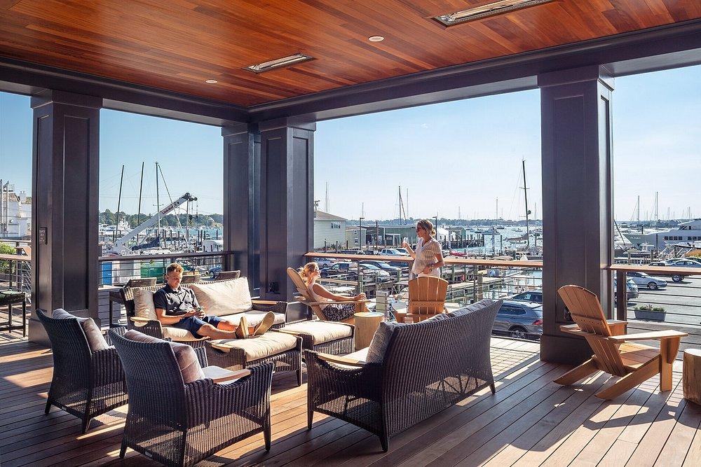 Outdoor patio with people relaxing, overlooking a marina on a sunny day.