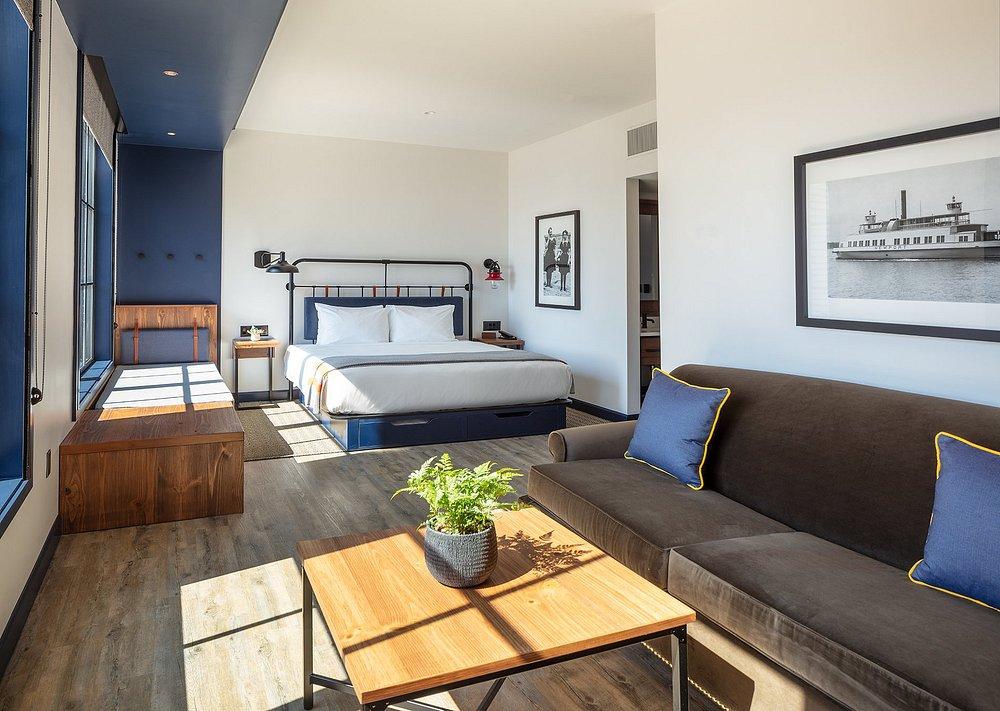 Modern hotel room with large bed, gray sofa, and wooden table with plant decor.