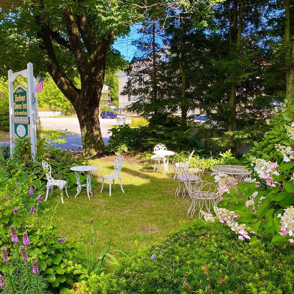Small garden with white chairs, tables amidst lush greenery and flowers, sunny day.