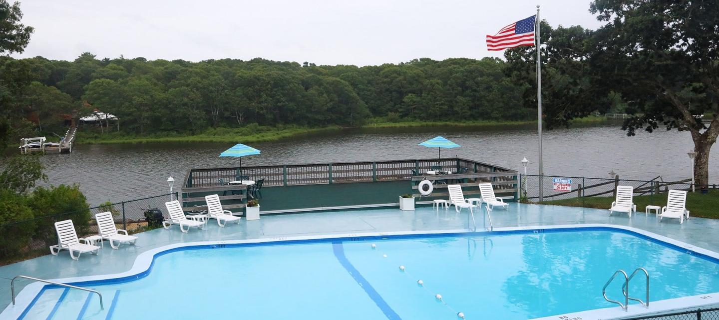 Outdoor pool with white chairs, overlooking a lake and lush green trees. American flag nearby.