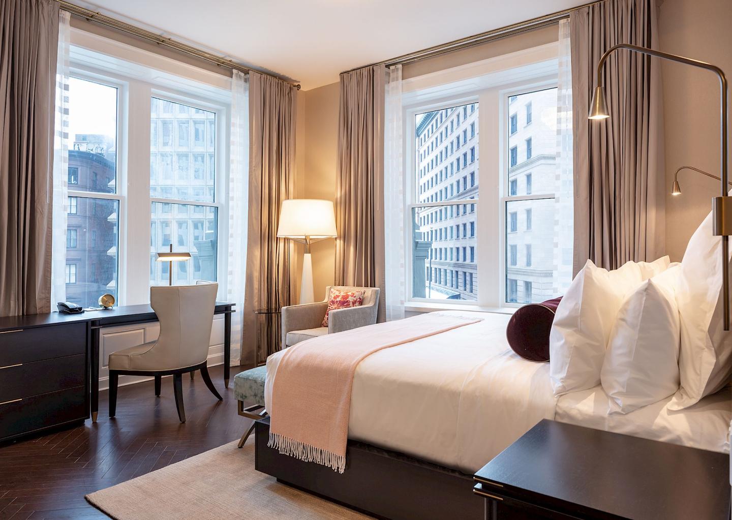 Cozy hotel room with large windows, bed, desk, and city view.