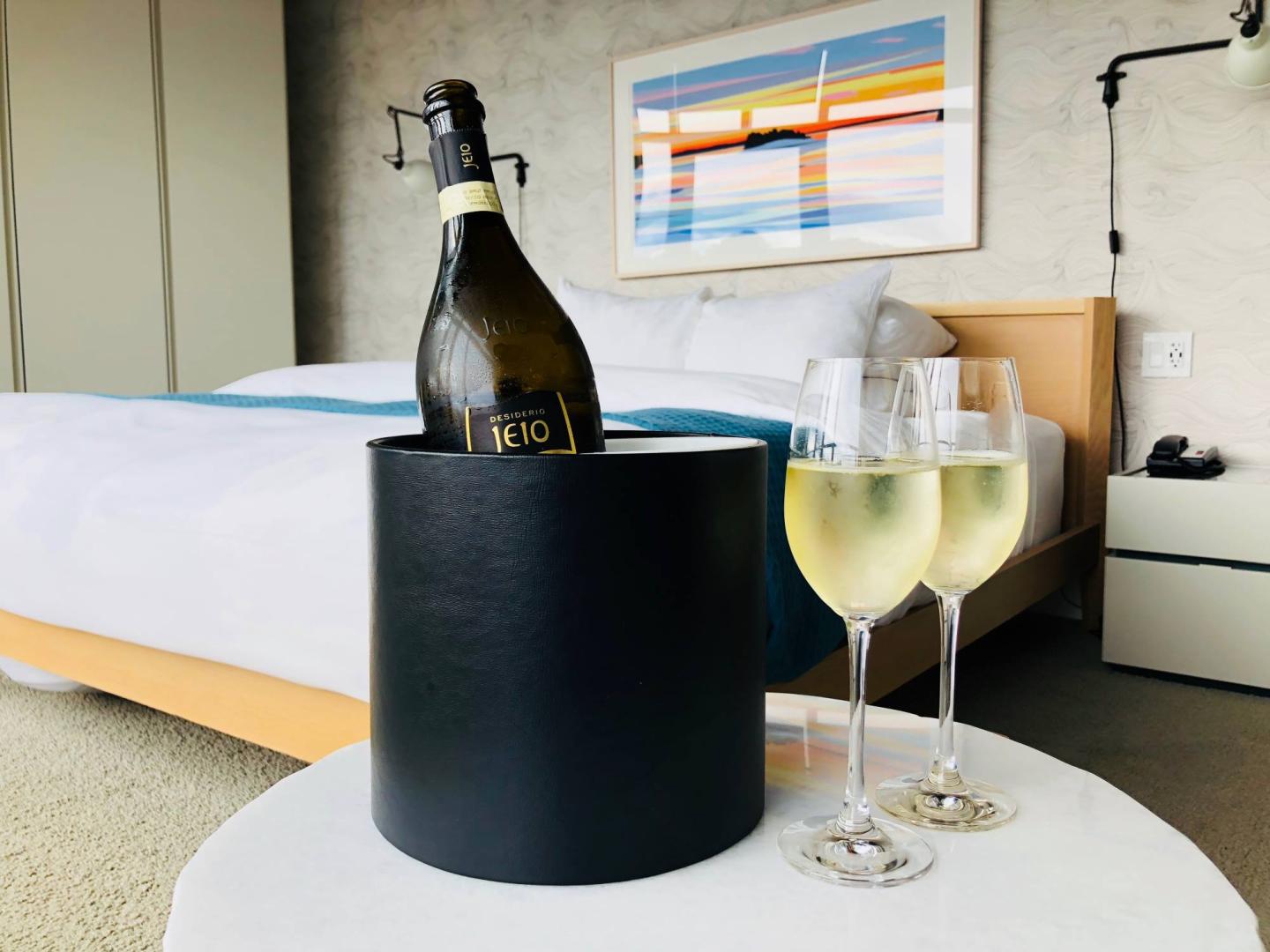 Wine bottle in an ice bucket with two glasses on a table in a modern bedroom.