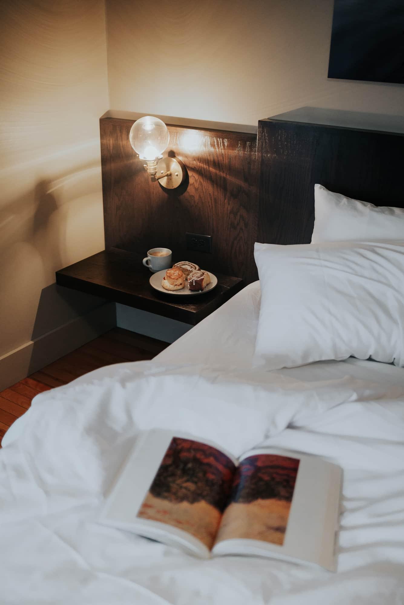 Cozy bed with open book, lit lamp, and snacks on bedside table.
