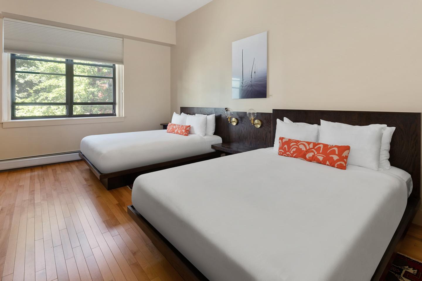 Two neatly made beds in a sunlit room with a large window.