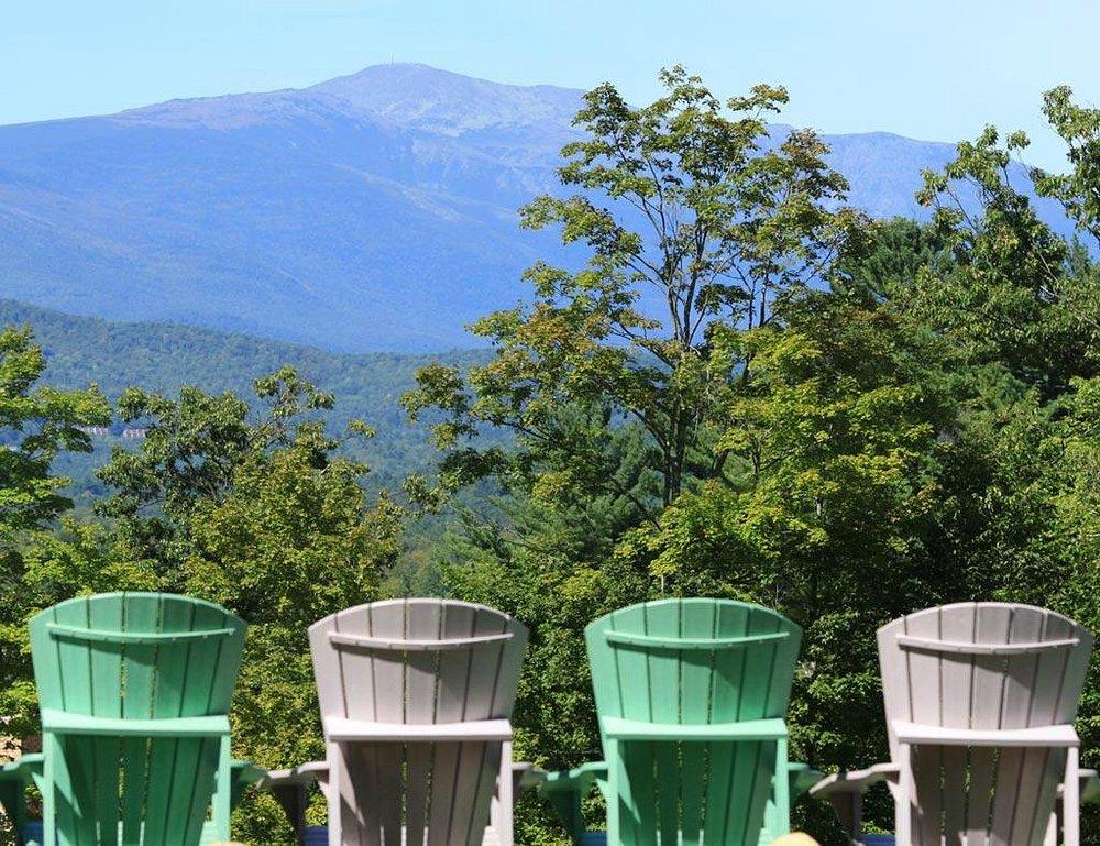 Four Adirondack chairs facing a mountain with lush trees in the foreground.