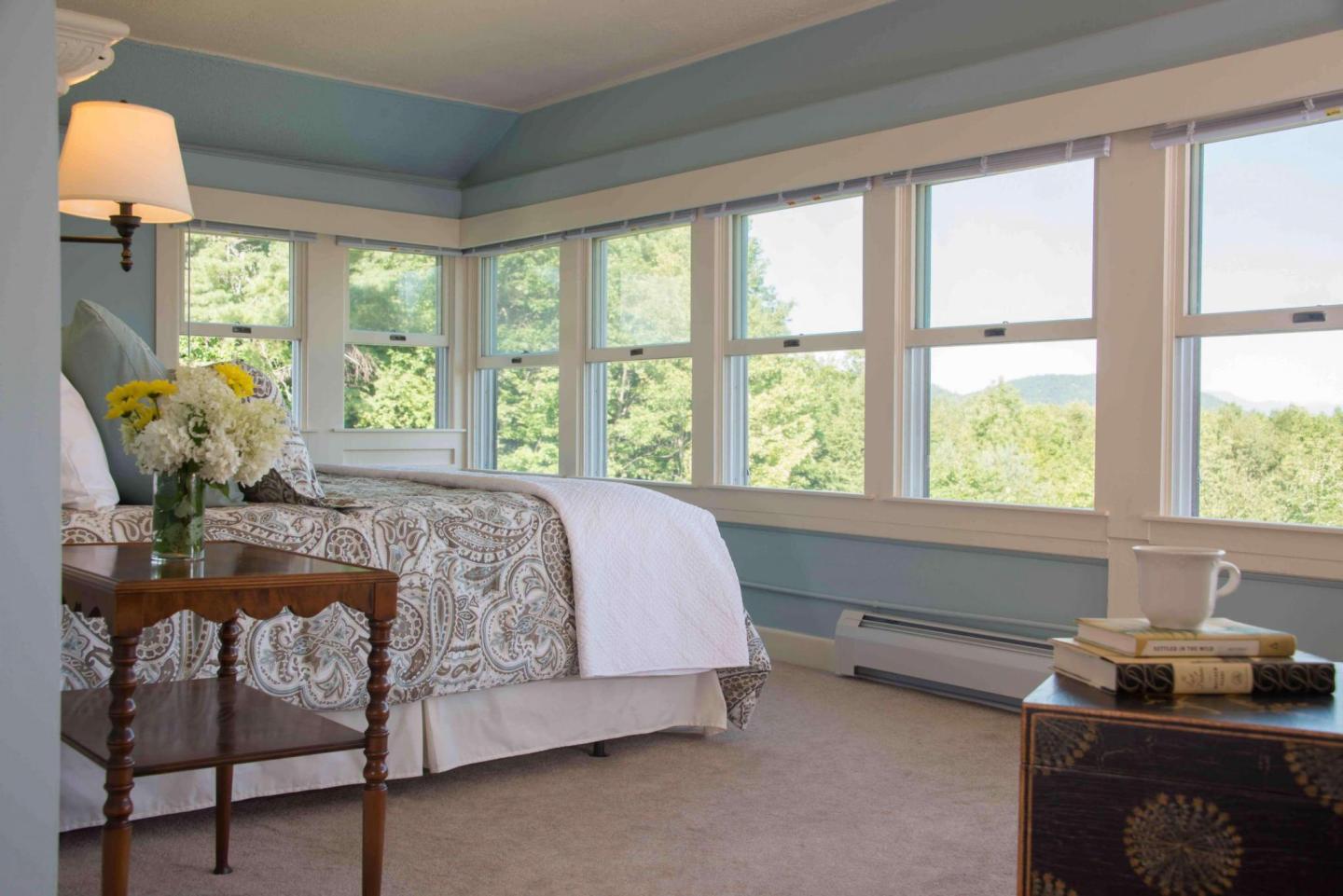 Cozy bedroom with large windows, floral bedding, and a side table with flowers.