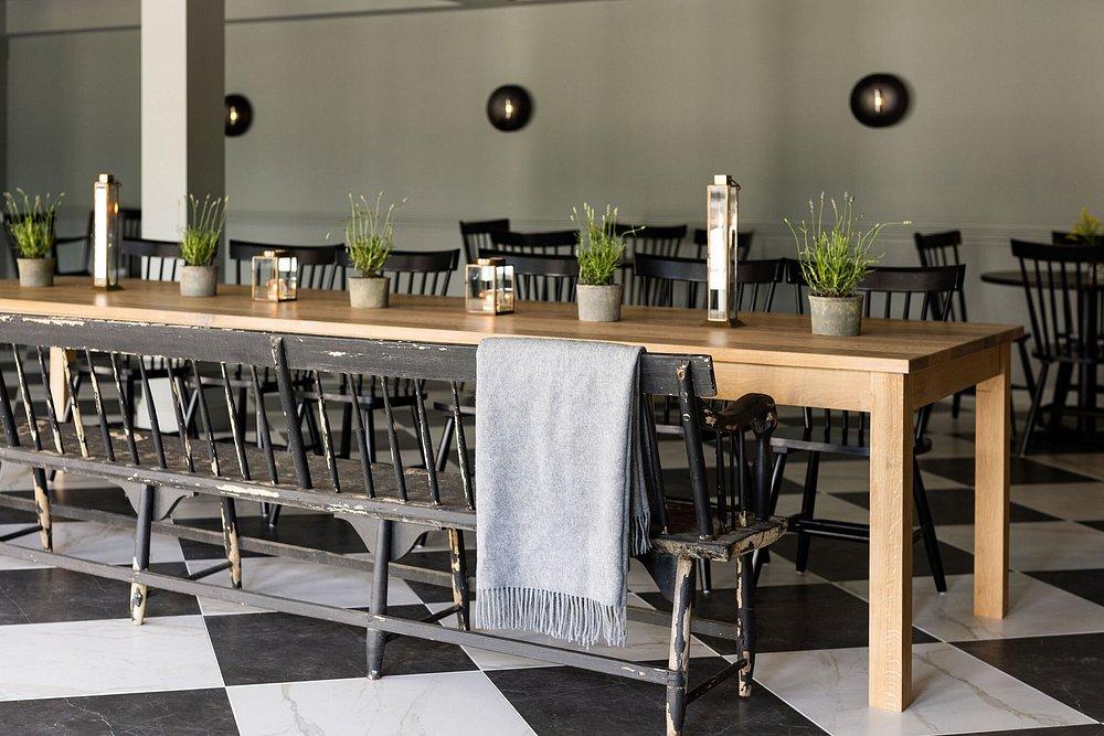 Long wooden table with plants, checkered floor, and black chairs.