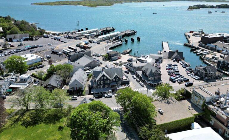 Harbor village with boats docked, buildings, trees, and a blue sea.